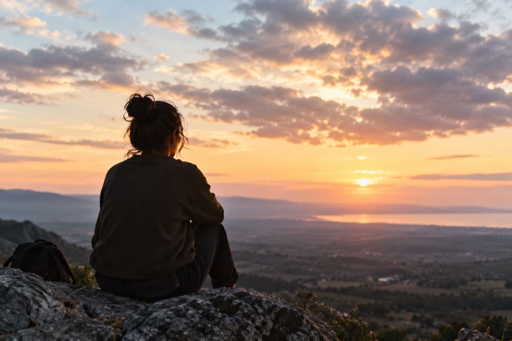 El mundo cambia cuando alguien se atreve a imaginarlo diferente Persona sentada de espaldas sobre unas rocas, contemplando un paisaje montañoso y el mar al atardecer, bajo un cielo cubierto de nubes iluminadas por una luz cálida.